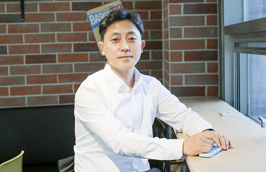 의자에 앉은 김상언 대표가 카메라를 응시하고 있다. 그는 테이블에 버사 슬레이트를 올려놓고 점필로 점자를 누르는 자세를 취하고 있다.