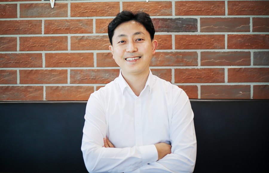 쇼파에 앉은 김상언 씨가 카메라를 응시하며 환하게 미소짓고 있다. 그는 하얀색 셔츠를 입고 있으며 팔짱을 끼고 있다.