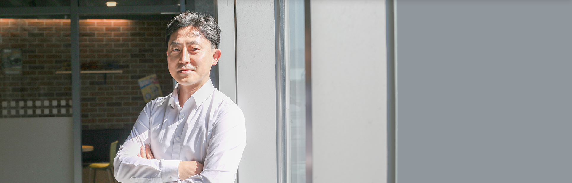 햇빛이 들어오는 유리 벽에 김상언 대표가 기대어 서 있다. 하얀 셔츠를 입은 그는 팔짱을 낀 채 카메라를 응시하며 미소짓고 있다.