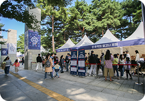 참여 행사 부스가 나란히 설치되어 있다. 그 앞에 수많은 사람이 줄을 서서 기다리고 있다. 사람들 주변에는 ‘추억의 달고나 체험, 한글을 뽑아라!’라는 문구와 함께 체험 시간표, 경품 안내 등을 적어놓은 입간판이 놓여있다. 멀리 기둥에는 ‘고마워 한글’이라고 적힌 포스터가 붙어있다.