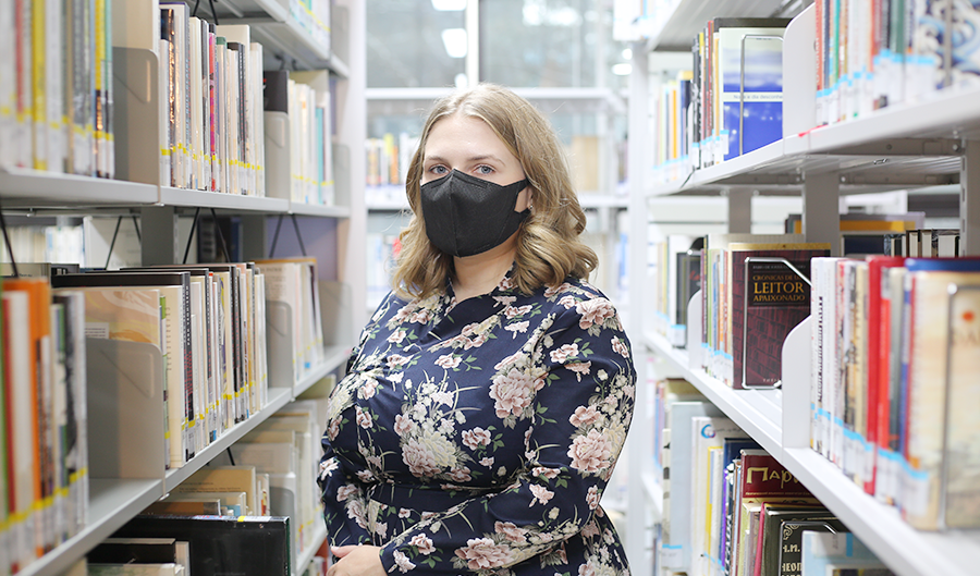 레르미트 쥬디트가 도서관 책꽂이 사이에 서 있다. 그녀는 검은 마스크를 착용했다. 그녀 양옆으로 책이 가득 꽂힌 책꽂이가 보인다.