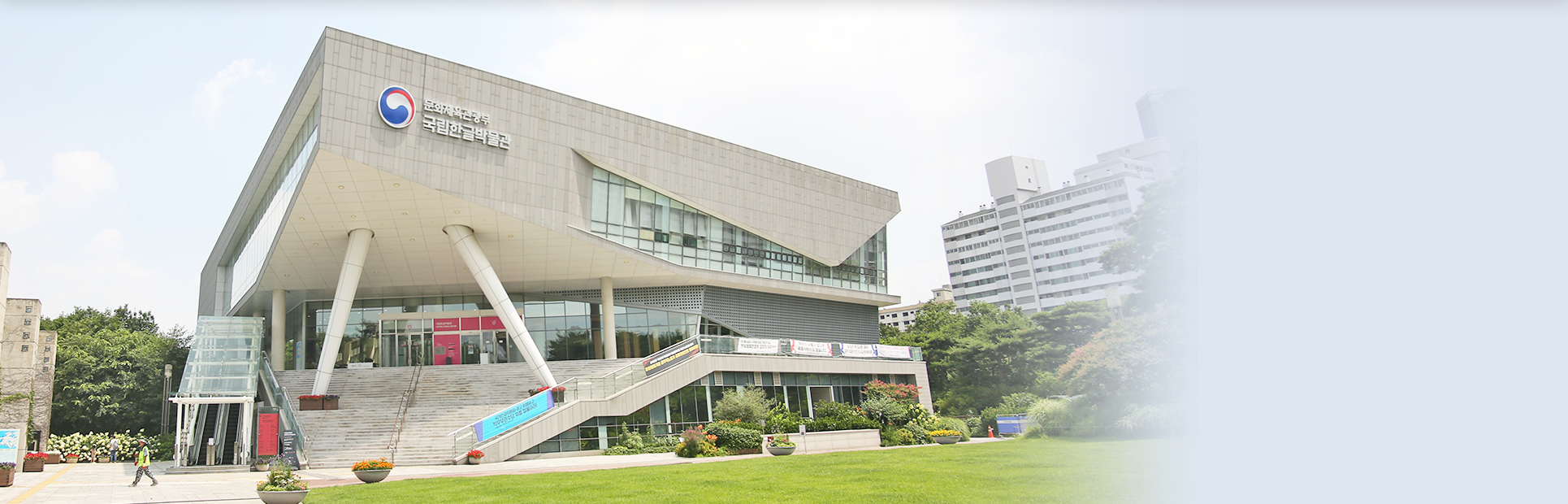 국립한글박물관 전경이다. 두 개의 기둥이 삼각형의 건물 머리를 받치고 있다. 주변으로는 녹색의 잔디와 녹음이 펼쳐져 있다.