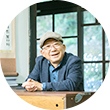 남색 정장과 파란색 셔츠를 입은 나태주 시인이 의자에 앉아 책상에 두 손을 모아 올리고 정면을 응시하고 있다. 나태주 시인은 안경과 모자를 썼으며 환하게 웃고 있다. 