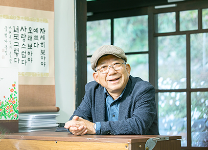 반갑습니다 기사 사진. 남색 정장과 파란색 셔츠를 입은 나태주 시인이 의자에 앉아 책상에 두 손을 모아 올리고 정면을 응시하고 있다. 나태주 시인은 안경과 모자를 썼으며 환하게 웃고 있다. 나태주 시인의 왼쪽엔 빨간 꽃과 풀잎 그림이 있고 그 뒤엔 흰색 종이들이 쌓여 있으며, 나태주 시인의 대표작인 「풀꽃」의 시구가 황토색 두루마기 형식의 종이에 담겨있다.