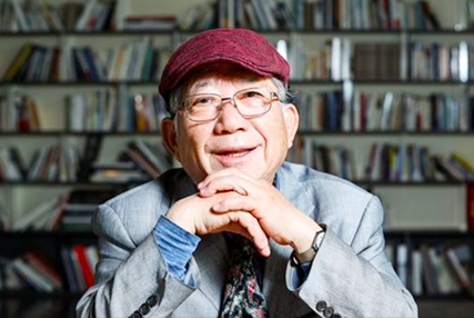 회색 정장을 입은 나태주 시인이 의자에 앉아 책상에 두 손을 올리고 턱을 괸 채로 정면을 응시하고 있다. 나태주 시인은 안경과 모자를 썼으며 그의 뒤에는 많은 책이 꽂혀있는 책장이 보인다.