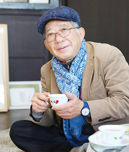 황토색 정장을 입고 파란색 목도리를 한 나태주 시인이 두 손으로 찻잔을 들고 정면을 응시하고 있다. 나태주 시인은 파란색 모자와 안경을 썼으며, 파란색 시계를 착용하고 있다.