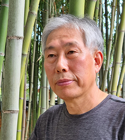 짙은 회색 반소매 티셔츠를 입은 이호신 화가가 대나무 숲에서 왼쪽을 바라보고 있다.