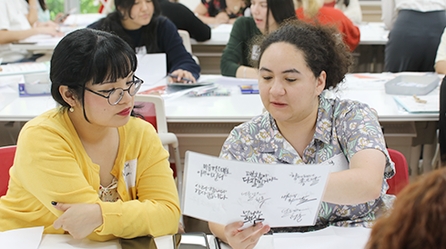 ‘손으로 꽃피우는 한글’ 교육 현장 사진이다. 외국인 수강생 2명이 의자에 앉아 한글 캘리그래피 작품을 보며 대화하고 있다.