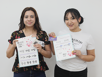 ‘손으로 꽃피우는 한글’ 교육 현장 미니 인터뷰 사진이다. 두 여성이 한글 캘러그래피 체험을 하며 쓴 종이를 들고 웃으며 정면을 바라보고 있다.