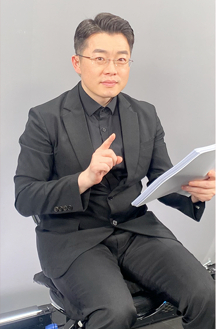 검은색 정장을 입고 안경을 쓴 김동호 수어 통역사가 비스듬히 앉아 정면을 응시하고 있다. 그는 오른손 검지를 피고 있으며 왼손엔 흰색 종이들을 들고 있다.