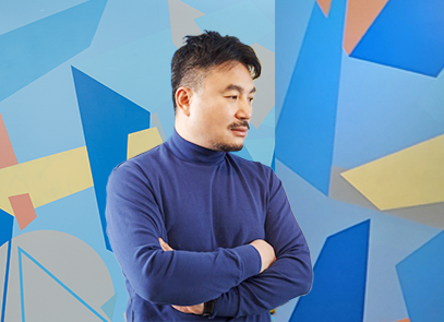 반갑습니다 기사 사진. 남색 옷을 입은 금보성 작가가 팔짱을 끼고 오른쪽 아래를 향해 바라보고 있다. 뒤에는 파랑, 주황, 노랑, 회색 등으로 한글 자음과 도형들이 곳곳에 그려져 있다.
