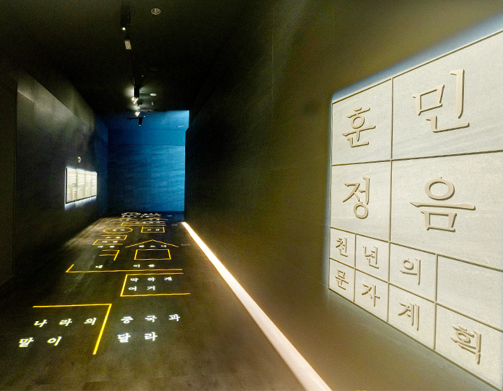 기획기사 기사 사진. ‘광복 78주년 기념 특별해설’ 상설전시실 입구 사진이다. 어두운 복도 아래 밝은 조명으로 훈민정음해례본의 내용이 적혀있으며, 벽에는 검은색 판 위에 설명들이 적혀있고 조명이 들어와 있다. 오른쪽엔 네모 한 칸씩 ‘훈민정음 천년의 문자’라고 적혀있다.