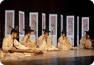전통음악집단 샛의 공연 사진이다. 전통의상과 갓을 쓴 다섯 명의 단원이 앉아서 제각각 전통 악기를 연주하고 있다. 무대 뒤에는 병풍 9개가 화면으로 띄워져 있다.