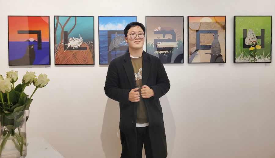 진관우 작가가 ‘ㄱ’, ‘ㄴ’ ‘ㄷ’, ‘ㄹ’, ‘ㅁ’, ‘ㅂ’ 등의 한글 자음이 그려져 있는 작품들 앞에 서서 카메라를 향해 포즈를 취하고 있다. 