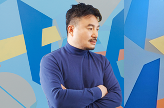 남색 옷을 입은 금보성 작가가 팔짱을 끼고 오른쪽 아래를 향해 바라보고 있다. 뒤에는 파랑, 주황, 노랑, 회색 등으로 한글 자음과 도형들이 곳곳에 그려져 있다.
