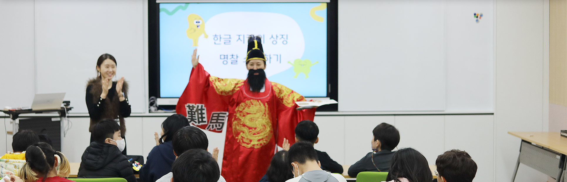 특수학급 학생들을 대상으로 한 한글문화 체험 교육 <세종의 친구, 한글 지킴이>의 시범 현장 사진이다. 임금님 복장을 한 강사가 왼손에 책을 들고 오른손은 옆으로 뻗으며 학생들을 가르치고 있고, 왼쪽엔 한 여성이 손뼉을 치고 있다.