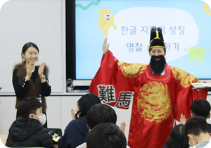 특수학급 학생들을 대상으로 한 한글문화 체험 교육 <세종의 친구, 한글 지킴이>의 시범 현장 사진이다. 임금님 복장을 한 강사가 왼손에 책을 들고 오른손은 옆으로 뻗으며 학생들을 가르치고 있고, 왼쪽엔 한 여성이 손뼉을 치고 있다.