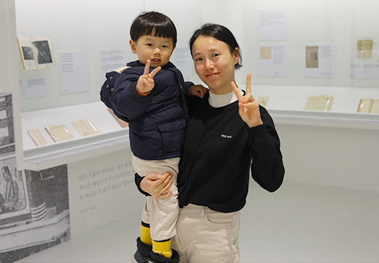 한글 노래 『한양가』를 중심 주제로 한 기획특별전 <서울 구경 가자스라, 한양가> 전시 현장 미니 인터뷰 사진이다. 한 여성이 남자 아이를 안고, 둘 모두 손가락을 이용해 ‘v’모양을 그리고 있다.