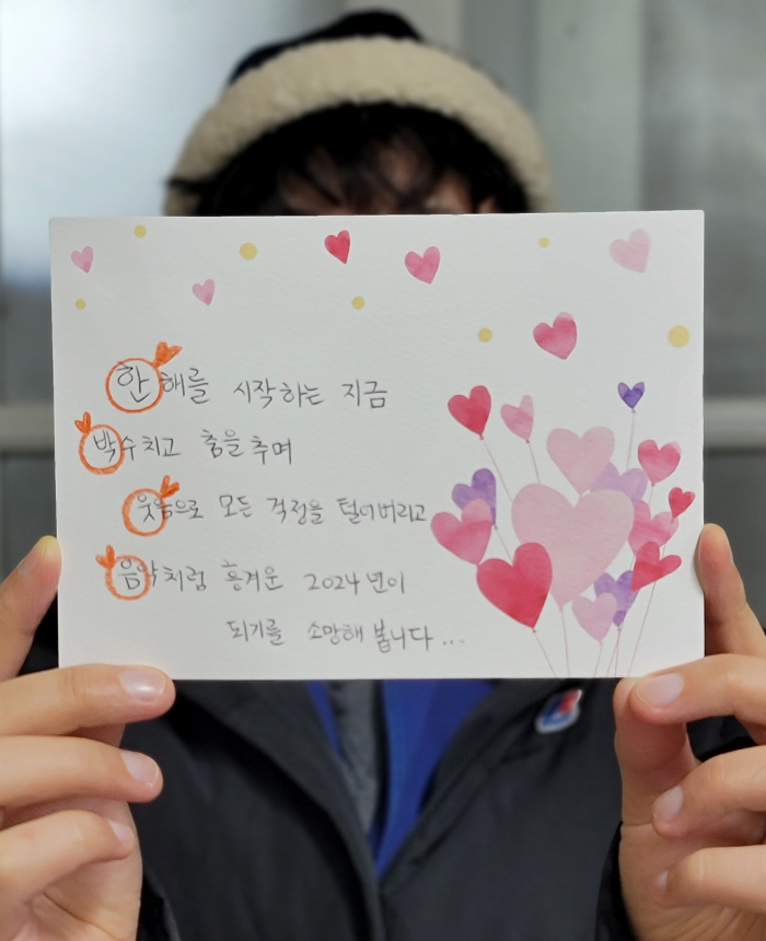 한 사람이 하트가 군데군데 그려진 카드를 들고 있다. ‘한: 한해를 시작하는 지금 박: 박수치고 춤을 추며 웃: 웃음으로 모든 걱정을 털어버리고 음: 음악처럼 흥겨운 2024년이 되길 소망해봅니다’