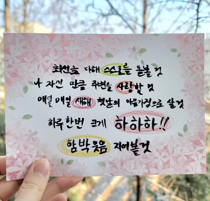 한 사람이 분홍색 꽃 배경의 카드를 들고 있다. ‘최선을 다해 스스로를 돌볼 것. 나 자신만큼 주변을 사랑할 것. 매일매일 새해 첫날의 마음가짐으로 살 것. 하루 한번 크게 하하하!! 함박웃음 지어볼 것.’