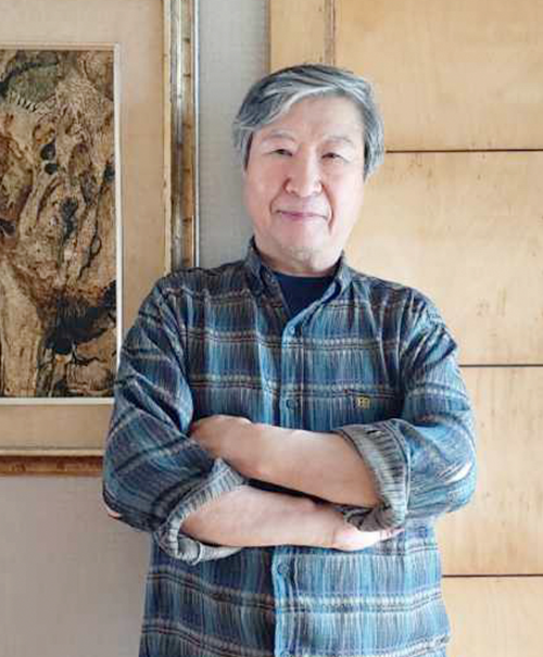 알록달록한 셔츠를 입은 김석희 교수가 팔짱을 낀 채 정면을 바라보고 있다. 그의 왼쪽 뒤엔 미술 작품이 걸려 있다. 