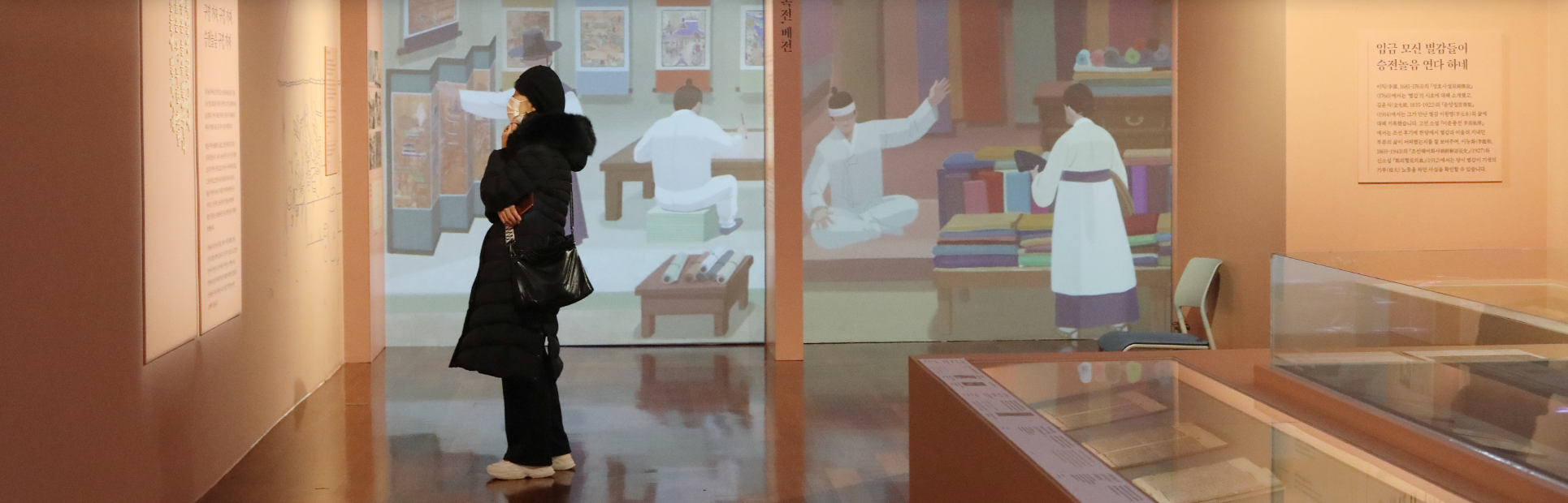 한글 노래 『한양가』를 중심 주제로 한 기획특별전 <서울 구경 가자스라, 한양가> 전시 현장 사진이다. 한 여성이 팔짱을 끼고 턱에 손을 올리며 벽에 있는 전시작품을 관람하고 있다. 그녀 뒤 스크린에는 조선시대를 나타내는 그림들이 비추어지고 있다. 