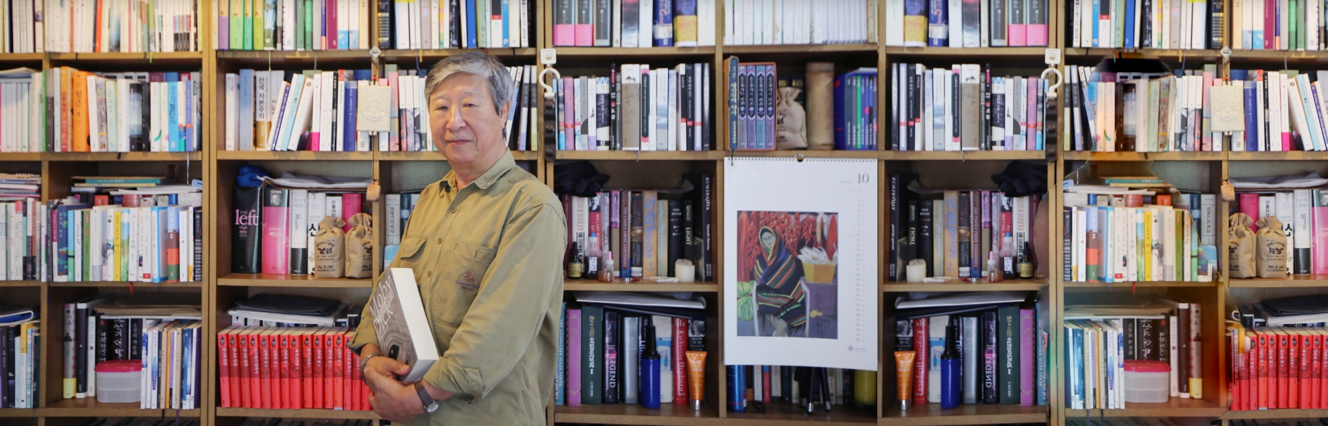김석희 번역가가 양손을 모아 도서 『모비 딕』을 들고 측면으로 비스듬히 서 정면을 바라보고 있다. 그의 뒤엔 책들이 빼곡하게 꽂혀있는 책장들로 꽉 차 있다.
