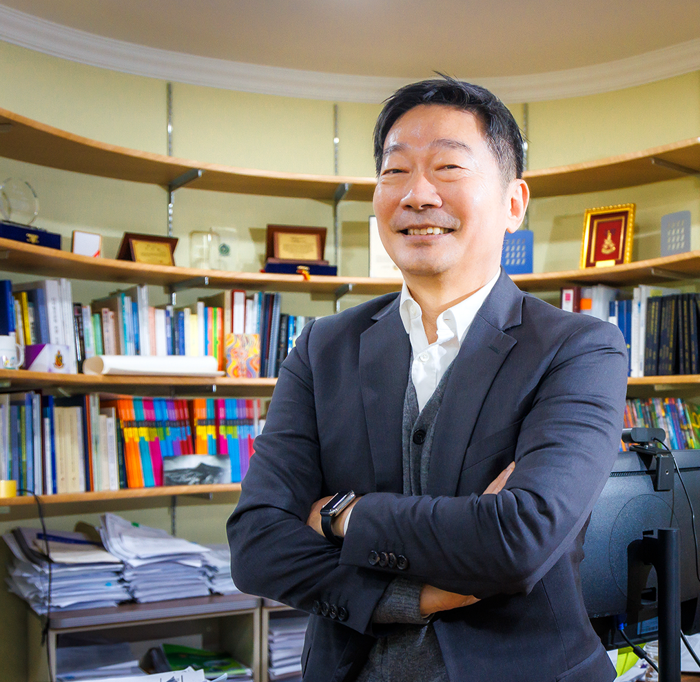 정장을 입은 김중섭 교수가 팔짱을 낀 상태로 비스듬히 서 정면을 보고 환하게 웃고 있다. 그의 뒤에는 컴퓨터 모니터와 빼곡하게 꽂혀있는 책장의 책들, 상패 등이 보인다.