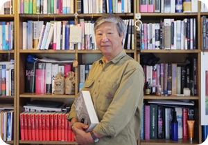 김석희 번역가가 양손을 모아 도서 『모비 딕』을 들고 측면으로 비스듬히 서 정면을 바라보고 있다. 그의 뒤엔 책들이 빼곡하게 꽂혀있는 책장들로 꽉 차 있다