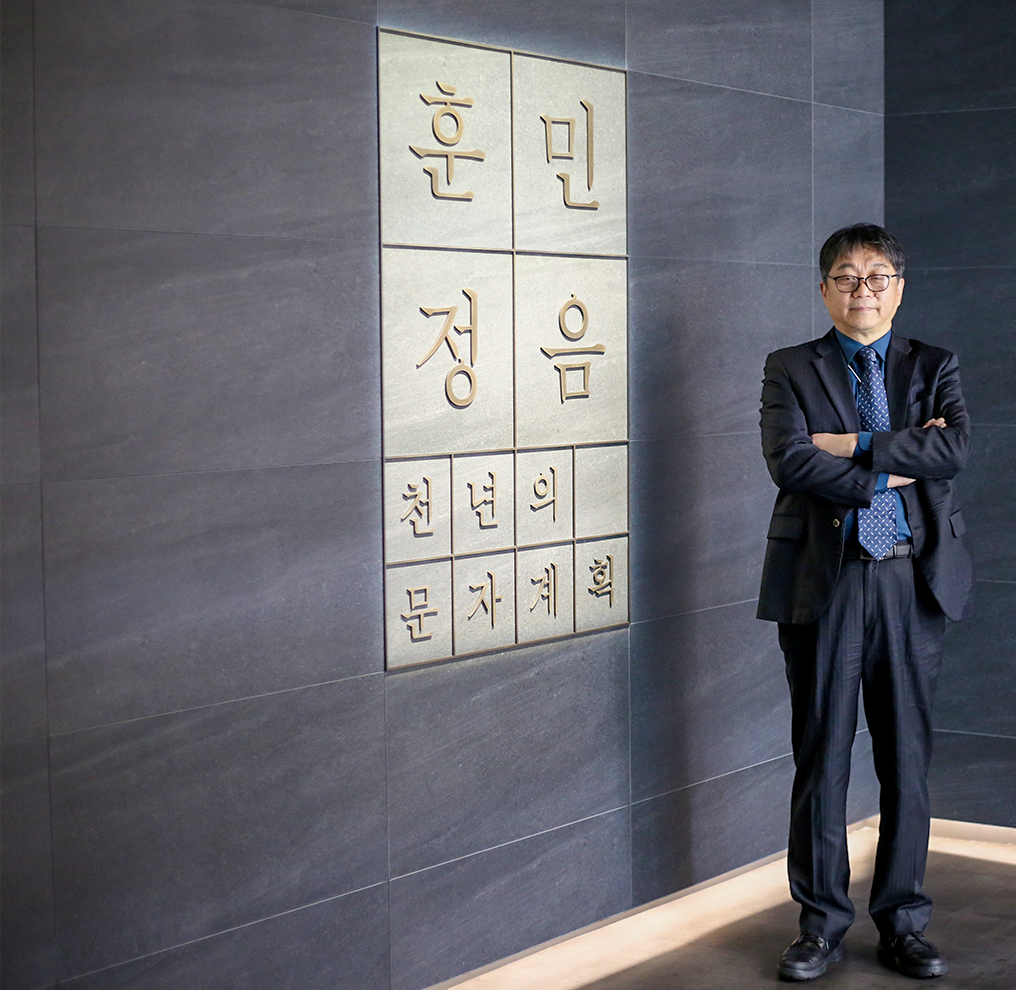 정장을 입고 안경을 쓴 김일환 관장이 팔짱을 낀 채 미소를 지으며 정면을 보고 서 있다. 벽에는 여러 개의 네모 칸이 붙어있고 각 네모 칸 안에는 '훈민정음 천년의 문자 계획'이라는 글이 글자 하나씩 적혀있다.