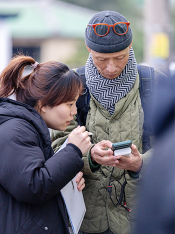 휴대전화를 보고 있는 남자와 여자 사진이 있다. 여자는 갈색 머리를 하나로 묶은 채로 남자가 들고 있는 휴대전화를 보고 있다. 남자는 회색의 모자 위에 주황색 안경을 걸친 차림으로 휴대전화를 들고 내용을 확인하고 있다. 