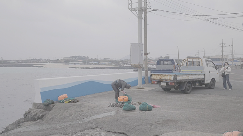 안개가 낀 것 같은 흐린 배경의 사진이다. 왼쪽에 바다가 있고, 가운데에 한 사람이 허리를 굽히고 서 있다. 그 옆으로는 트럭이 한 대 서 있다. 가장 왼쪽에는 여자가 한 명 서 있다. 