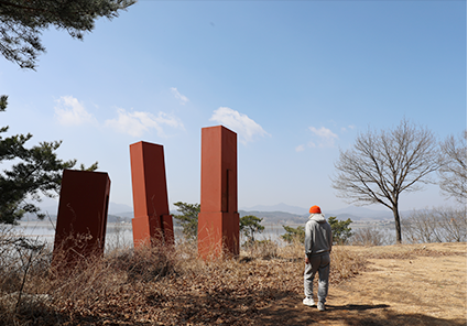 바다를 배경으로 서 있는 한창조 작가의 뒷모습이다. 한창조 작가의 앞에 빨간색 기둥 모양의 작품이 서 있다. 그 옆에는 나무 두 그루가 서 있다.