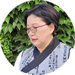 담쟁이덩굴 앞에 권숙희 낭송가가 자신이 들고 있는 두루마리를 바라보며 서 있다. 권숙희 낭송가는 옛한글이 프린트된 저고리를 입고 있으며 안경을 쓰고 있다.