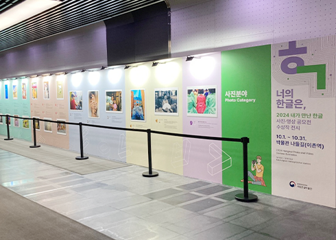 이촌역 박물관 나들길 전시 전경 사진이다. 긴 복도를 따라 ‘제4회 내가 만난 한글 사진영상 공모전 전시’ 사진 부분 수상작이 전시되어 있다. 벽면을 따라 여러 개의 사진이 일정한 간격으로 배치되어 있고 각각의 사진 아래에는 작품 설명이 적혀 있다. 벽은 밝은 색상으로 칠해져 있으며, 사진 위에는 개별 작품을 비추는 조명이 달려 있다. 작품이 전시된 벽면 앞쪽에는 안전을 위해 검은색 벨트가 설치되어 있다.