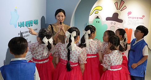 한글놀이터 순회전 ‘나의 특별한 하루’ 개막식 행사 현장 사진이다. 큐레이터의 전시 설명을 듣고 있는 아이들이 있다. 여자아이들은 꽃무늬 상의와 분홍색 한복 치마를 입고 있고, 남자아이들은 파란색 한복 조끼를 입고 있다. 큐레이터와 아이들은‘나의 특별한 하루‘라는 문구가 적혀 있는 밝은 파란색 전시 벽면 앞에 서 있다.