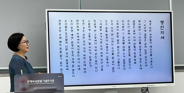 훈민정음 해례본 마지막에 실린 ‘정인지서’를 한글 가사체로 써서 강의하는 권숙희 낭송가의 사진이다. 사진 오른쪽에 ‘국제보상운동기념도서관’이라고 쓰여 있는 발표대 뒤에서 초록색 상의를 입고 안경을 쓴 권숙희 낭송가가 오른쪽을 바라보고 있다. 그의 옆에는 큰 모니터가 있고 그 안에 ‘정인지서’가 한글 가사체로 풀이되어 있다. 