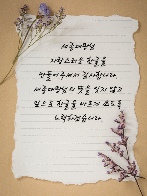 이벤트 예시로 쓰인 손 편지(글) 사진이다. 갈색 바탕에 흰색 편지지가 놓여있다. 편지지 안에는 ‘세종대왕님 자랑스러운 한글을 만들어주셔서 감사합니다. 세종대왕님의 뜻을 잊지 않고 앞으로 한글을 바르게 쓰도록 노력하겠습니다.’라고 적혀있다. 편지지 왼쪽 상단과 오른쪽 하단에는 드라이플라워가 각각 놓여 있다.