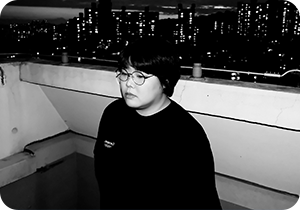 김영선 디자이너를 담은 흑백 사진이다. 앞머리가 있는 짧은 머리를 한 김영선 디자이너는 둥근 테의 안경을 썼고, 검은색 옷을 입고 있다. 시선은 왼쪽 아래를 향하고 있다. 그녀의 뒤로는 건물 옥상 난간이 있고 난간 바깥쪽으로는 빛을 뿜어내고 있는 건물이 있다.