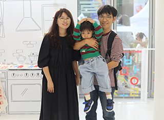 최정희, 강선우, 강태철 가족사진이다. 검은색 원피스를 입고 있는 최정희 씨는 카메라를 바라보며 웃고 있다. 그 옆에 아들 강선우 군을 안고 웃고 있는 강태철 씨가 있다. 강선우 군은 양손을 머리 위에 얹고 있다.
