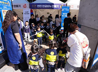 ‘2024 한글주간’ 현장 모습이다. 남색 유치원복을 입은 여러 명의 어린이가 모여서 체험을 해보고 있다. 어린이들 주변에는 선생님 두 분이 서서 지켜보고 있다. 체험하는 어린이들 뒤로 ‘천연 한글비누 만들기’ 체험 부스가 있다.