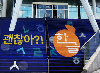 ‘2024 한글주간’ 현장 모습이다. 계단에 ‘괜찮아?! 한글’ 로고가 랩핑되어 있다. 왼쪽 계단에는 남색 바탕에 노란색으로 ‘괜찮아?!’라고 적혀 있고, 주변에 한글 자음이 그려져 있다.
                                    오른쪽 계단에는 똑같이 남색 바탕에 주황색 원이 그려져 있고, 그 안에 ‘한글’이라고 적혀 있다. 로고 주위로 한글 자음이 그려져 있다.