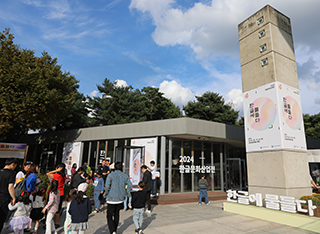 ‘2024 한글주간’ 현장 모습이다. ‘2024 한글문화산업전’이 열리고 있는 국립한글박물관 별관 입구 앞에 사람들이 모여있다. 별관의 오른쪽에는 커다란 직사각형 콘크리트 기둥이 서 있고, 기둥에는 ‘한글에 물들다’라고 적혀 있는 두 개의 큰 흰색 포스터가 붙여져 있다. 콘크리트 기둥 앞에는 ‘한글에 물들다’라는 문구가 적힌 조형물이 있다.