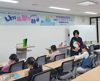 ‘한글로 뭉치고 펼치고’ 교육 현장 사진이다. 두 줄로 된 책상 앞에 4명의 여자아이가 앉아 폼포드에 한글 자음과 모음을 그리는 작업을 하고 있다. 피카소의 작품이 그려진 앞치마를 입고 있는 교육 담당자 정선희 작가는 그림을 그리고 있는 학생을 내려다보고 있다.