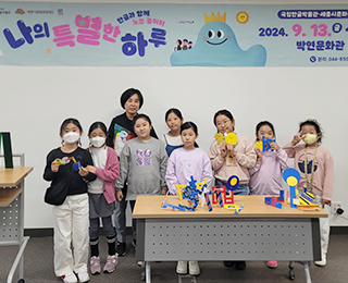 ‘한글로 뭉치고 펼치고’ 교육 현장 사진이다. 정선희 작가와 수업을 받은 아이들이 함께 모여 카메라를 바라보고 있다. 몇몇 아이들은 자신들이 만든 작품을 들고 웃고 있다. 아이들 앞에 있는 테이블 위에는 아이들이 직접 만든 한글 입체 조형물이 세워져 있다. 정선희 작가와 아이들 뒤쪽 벽에는 ‘나의 특별한 하루’ 행사명과 일정이 적혀 있는 현수막이 걸려있다.