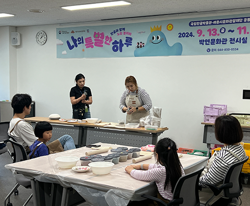‘한글로 뭉치고 펼치고’ 교육 현장 사진이다. 한쪽 벽에 ‘나의 특별한 하루’ 행사명과 일정이 적혀 있는 현수막이 걸려있다. 사진 중앙에는 큰 테이블이 있고 그 위에는 도예 도구와 재료가 놓여있다. 테이블 앞에는 2명의 여자아이와 각각 아이들의 보호자가 앉아 있다. 이들은 베이지색 앞치마를 입은 러블리킴 작가가 도자기 만들기 시범을 보이는 걸 쳐다보고 있다. 러블리킴 작가 앞에는 테이블이 있고, 그 위에 도예 재료와 도구가 놓여있다.
