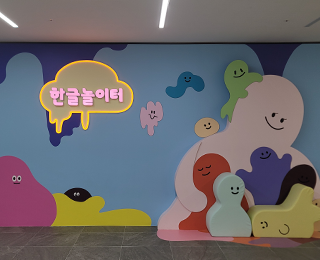한글놀이터 순회전 ‘나의 특별한 하루’ 전경 사진이다. 밝고 아기자기한 디자인의 벽면이 있다. 벽면 중앙에는 노란색 테두리로 녹아내리는 구름이 그려져 있으며, 그 안에 ‘한글놀이터’라고 분홍색으로 크게 적혀 있다. 초록색, 분홍색, 노란색 등 다채로운 색깔의 캐릭터들이 벽면에 그려져 있고, 그 앞에 조형물로도 있다.
