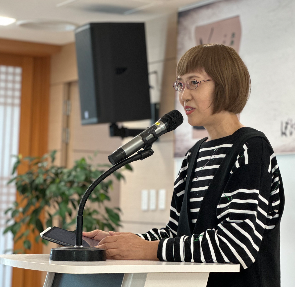 반갑습니다 사진. 시미즈 치사코 번역가가 마이크 앞에 서서 말하고 있다. 시미즈 치사코 번역가는 턱선 위로 올라오는 짧은 갈색 단발에 안경을 쓰고 있고 줄무늬 티셔츠를 입고 있다. 두 손으로 아이패드를 만지고 있다.