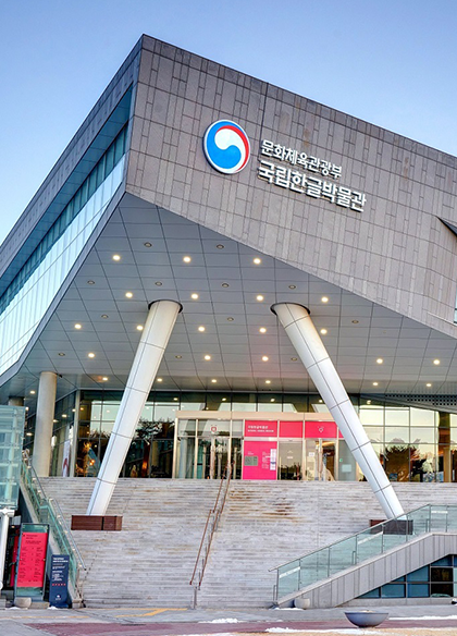 기획 기사 사진. 국립한글박물관의 전면 외관 사진이다. 건물의 상단에는 ‘문화체육관광부 국립한글박물관’이라는 한글과 로고가 있다. 건물의 외관은 밝은 회색 계열이며, 현대적인 건축 양식을 가지고 있다. 사진의 중앙에는 건물의 출입구로 통하는 넓은 계단이 있다.