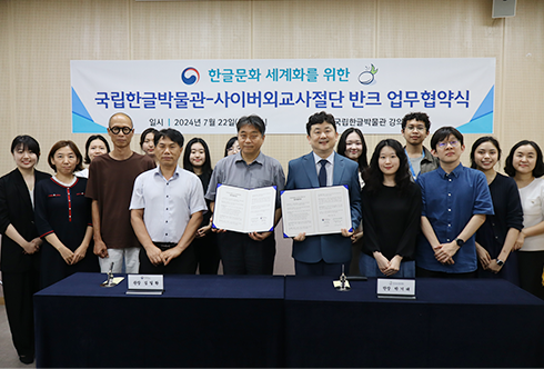 국립한글박물관과 사이버외교사절단 반크의 업무협약식 기념사진이다. 가운데 두 명은 협약서를 들고 미소 짓고 있으며, 그들의 주변에는 국립한글박물관과 사이버외교사절단 반크의 직원들이 서 있다. 사진 뒤쪽에는 ‘한글문화 세계화를 위한 국립한글박물관-사이버외교사절단 반크 업무 협약식’이 적혀 있는 커다란 현수막이 걸려있다.